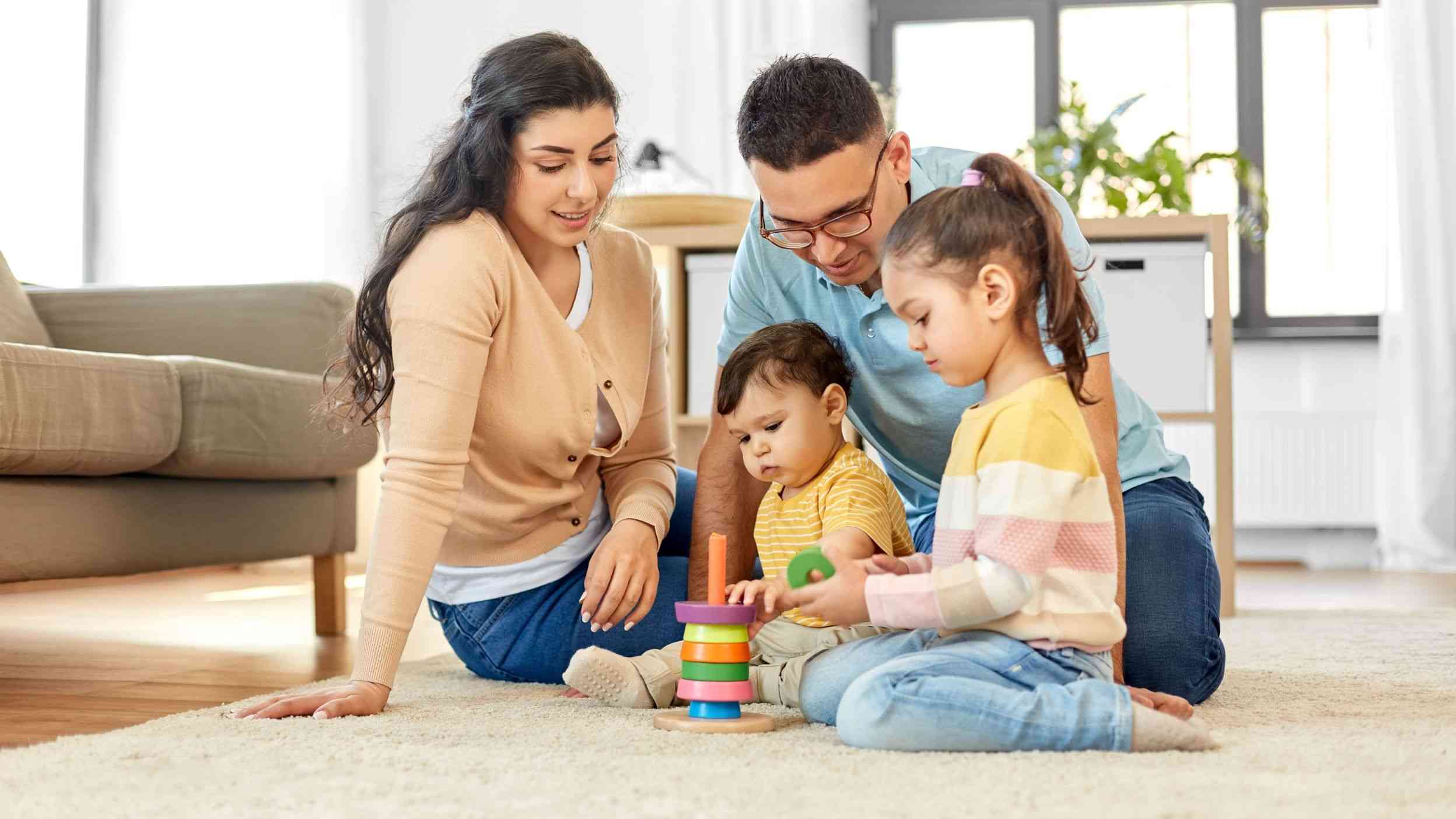 A familly sitting closely on a rug while the toddler builds a toy tower