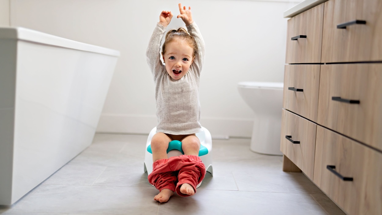 An excited toddler holding up her arms while sitting on a potty