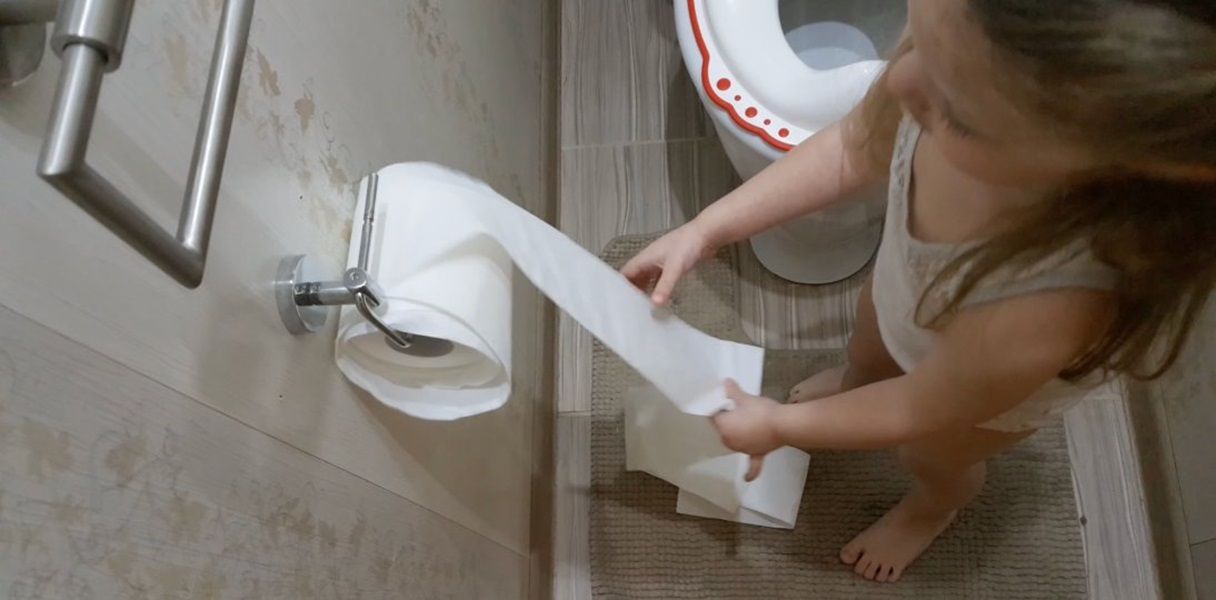 A girl standing next to the toilet while rolling down toilet paper