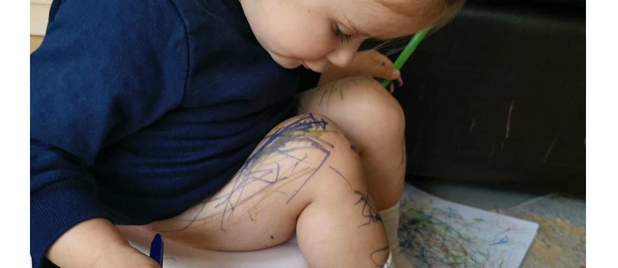 A toddler sitting on the toilet and scribbling on her leg with coloured pens