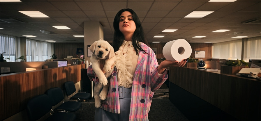 A confident girl wearing sunglasses, standing beside a puppy while holding a toilet roll.