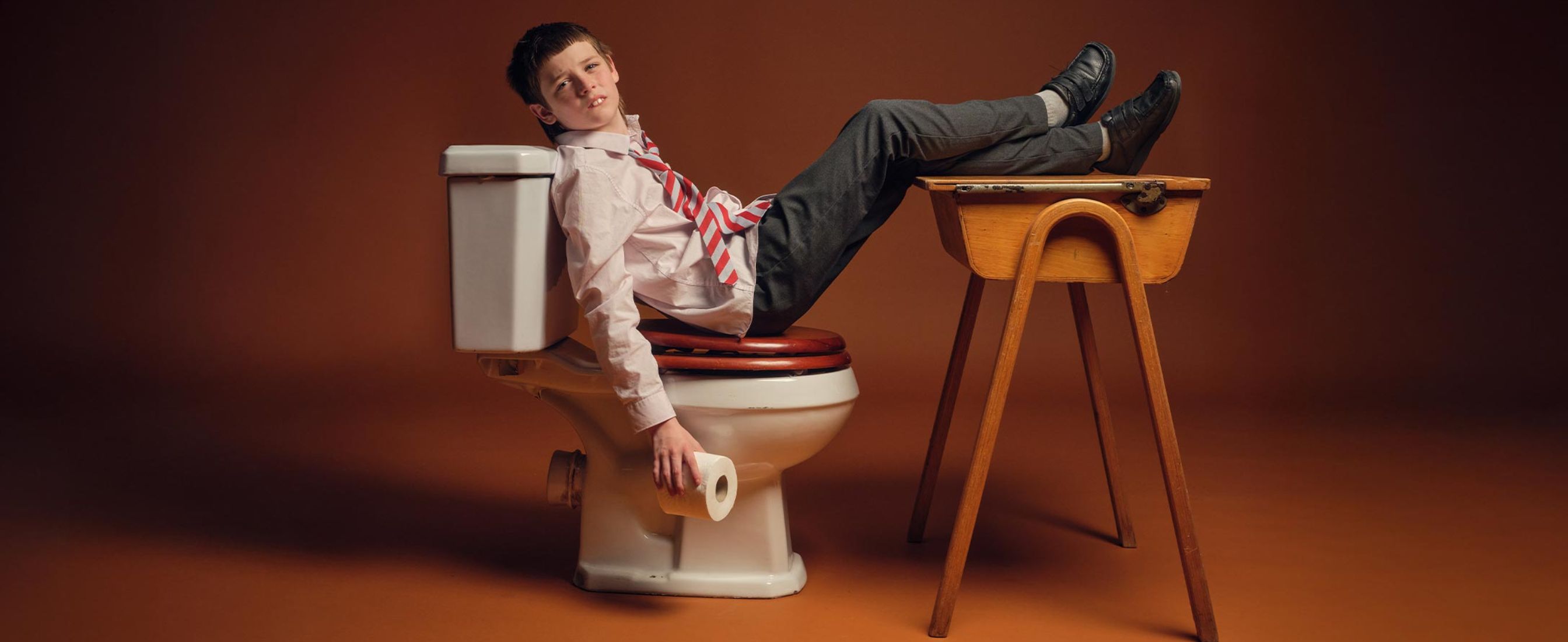 A confident school boy sitting on a toilet with his feet propped up on school books
