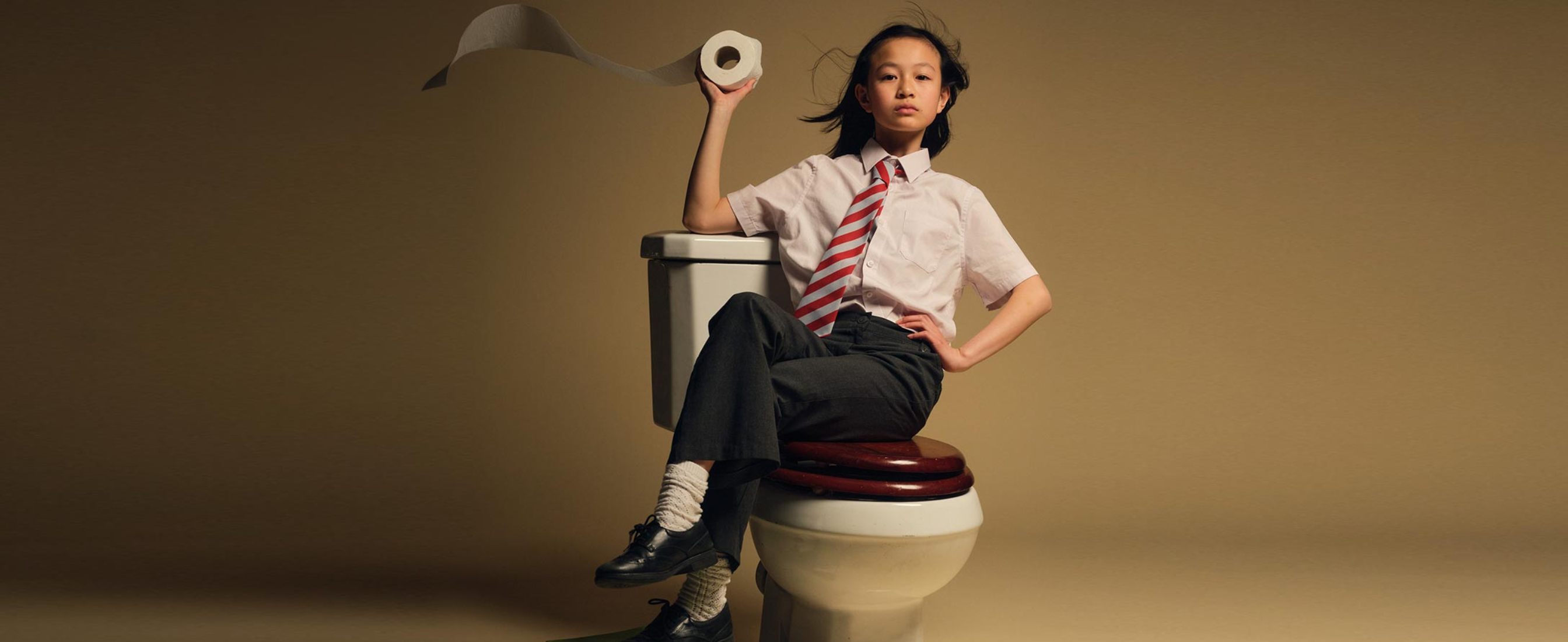 A confident school boy sitting on a toilet with his feet propped up on school books